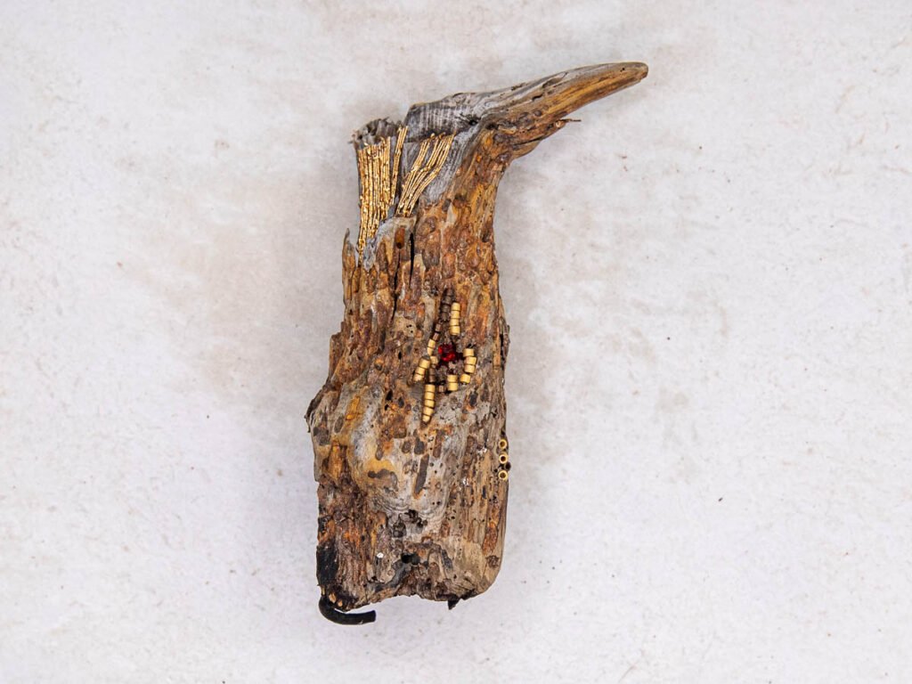 Fragment de bois sculpté avec incrustations de perles cylindriques dorées et rouges, fond minéral pâle.